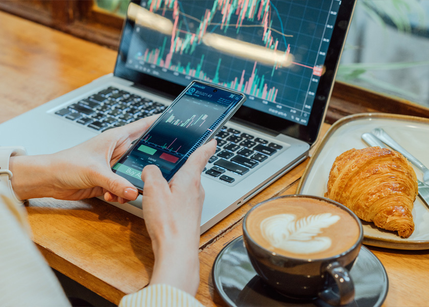 Image of woman holding phone with stocks on screen and her laptop on the desk with stocks on screen. There's also coffee and a croissant on the table.